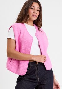 Pink fleece vest with a snap button closure, round neckline, and side pockets, paired with a white shirt and dark jeans.