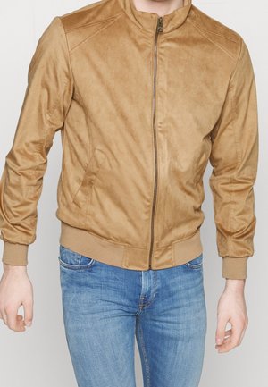 Beige Wildleder-Bomberjacke mit Frontreißverschluss an einem Mann mit blauen Jeans, von Hals bis Hüfte vor schlichtem Hintergrund gezeigt.