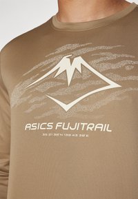 Hnedé športové tričko s bielym logom hory a textom "ASICS FUJI TRAIL" nad súradnicami, vyrobené z hladkého, vlhkosť odvádzajúceho materiálu.