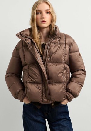 Young woman with long blonde hair wearing a brown padded jacket with high collar and dark blue jeans, hands in pockets, neutral background.