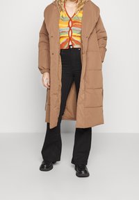 Hnědý péřový kabát s kapucí, vyrobený z prošívaného materiálu a zapínající na patentky. Nosený přes multikolorový vzorovaný top a černé zvonové kalhoty.