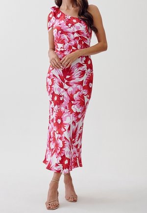 Vrouw draagt een één-schouder, enkellange jurk met groot rood en roze bloemenpatroon en nude sandalen met bandjes en hakken.