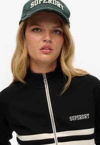 Veste noire zippée avec des bandes blanches, arborant un logo brodé "SUPERDRY". Accompagnée d'une casquette de baseball verte avec des lettres blanches.