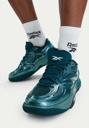 Persona che indossa scarpe da ginnastica Reebok turchesi e calzini Reebok bianchi alti, in piedi su una superficie bianca con un piede leggermente sollevato.