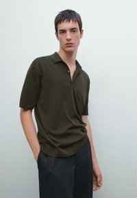 Polo a maniche corte di colore verde oliva, realizzato in tessuto morbido, con colletto e patta con tre bottoni, abbinato a pantaloni scuri.