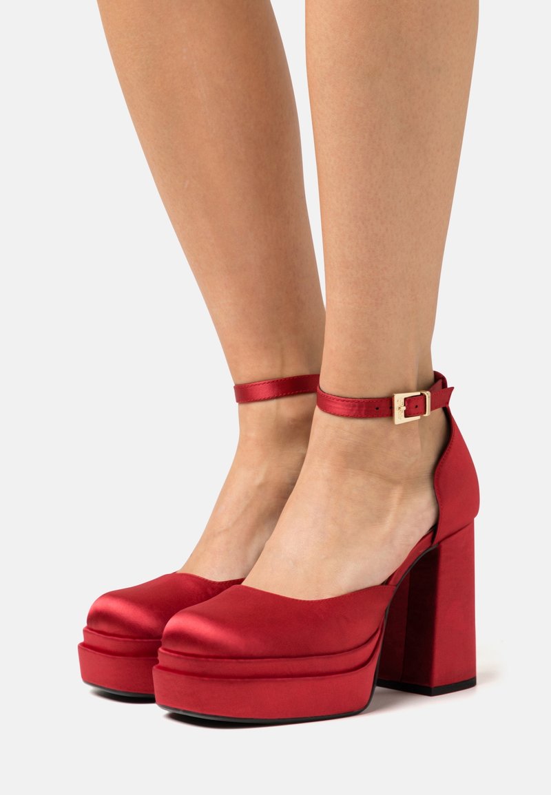 Pieds chaussés de talons compensés en satin rouge avec brides à la cheville et boucle dorée, présentés sur un fond clair uni.
