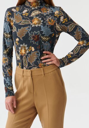 Kvinde iført en langærmet højhalset bluse med blomsterprint og højtaljede camel-farvede skræddersyede bukser, med hånden hvilende på hoften.