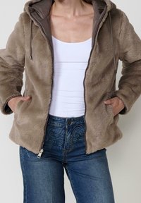 Personne portant une veste zippée taupe duveteuse par-dessus un débardeur blanc et un jean bleu à motifs, mains dans les poches de la veste.