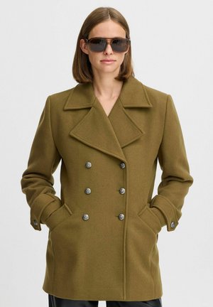 Manteau en laine vert olive avec un design croisé, grands revers, deux poches avant et boutons argentés. Le mannequin porte des lunettes de soleil oversize.