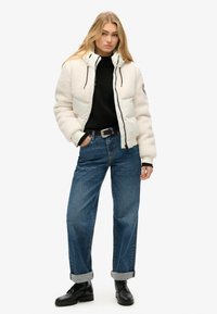 Veste en peluche blanche et crème avec des accents noirs, associée à un pull noir et un jean bleu taille haute. Des bottes noires à lacets viennent compléter le look.
