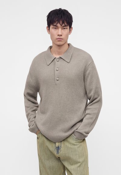Jeune homme aux cheveux courts noirs portant un pull en maille beige avec des boutons et un pantalon ample vert clair, debout les mains dans les poches.
