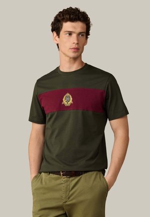 Mann mit lockigem Haar, der ein dunkelgrünes T-Shirt mit bordeauxrotem Streifen und goldenem Emblem trägt, kombiniert mit hellolivfarbenen Hosen und geflochtenem Gürtel.