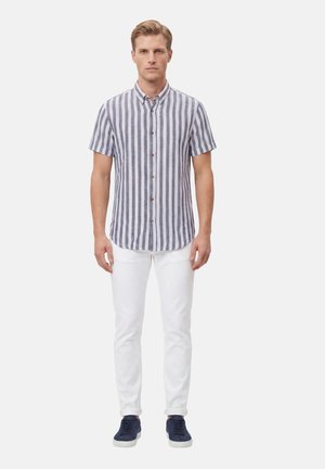 Uomo che indossa una camicia a righe verticali a maniche corte con bottoni, pantaloni bianchi e sneakers blu navy, in piedi rivolto verso il davanti su sfondo bianco.