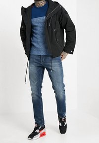 Schwarze Zip-Jacke mit Kapuze, blauer strukturierter Pullover, ausgewaschene blaue Jeans und schwarze Sportschuhe mit roten Akzenten.