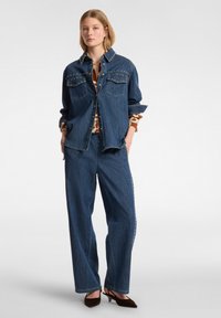 Jeansowa kurtka z ćwiekami, dwa kieszenie na piersi; połączona z szerokimi spodniami dżinsowymi. Model nosi wzorzystą koszulę pod kurtką.