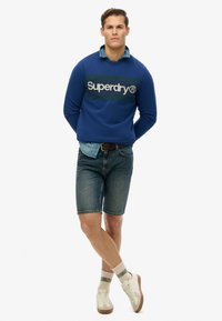 Niebieska bluza z białym logo "Superdry", w połączeniu z dżinsowymi szortami i białymi trampkami. Posiada warstwowy kołnierz dżinsowy oraz wzorzyste skarpetki.