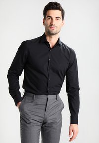Camicia da uomo nera a maniche lunghe con colletto classico; presenta una vestibilità slim e una chiusura con bottoni; indossata con pantaloni eleganti grigi.