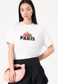 T-shirt en coton blanc avec un motif de framboises au-dessus du mot "PARIS" en lettres noires en gras. Associé à une jupe noire et un sac rose.