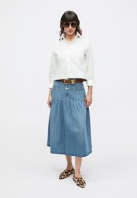 White cropped shirt, blue denim skirt with gathered detail, brown belt with gold buckle, and leopard print slip-on shoes. Model wearing sunglasses.