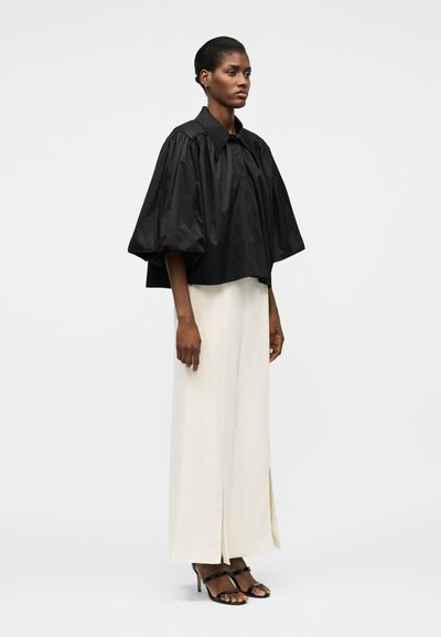 Femme debout portant un chemisier noir surdimensionné à manches ballon, un pantalon large crème avec des fentes à l'avant, et des sandales à talons ouvertes noires sur un fond uni.