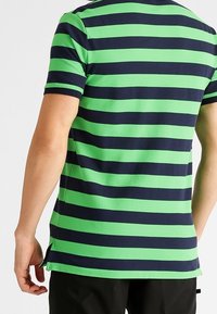 Kortärmad pikétröja med växlande gröna och marinblå horisontella ränder, tillverkad av ett mjukt, stretchigt tyg med sidslitsar.