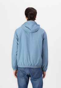Lyseblå hettejakke laget av lett materiale, med elastiske mansjetter og nederkant, vist fra baksiden sammen med casual blå jeans.