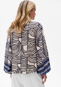 Geprinte blouse met een beige basis, voorzien van zwarte golfpatronen en blauwe geometrische accenten op de mouwen. Lichte stof, ontspannen pasvorm.