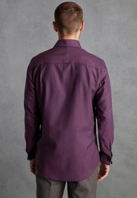 Mann mit dem Rücken zur Kamera, der ein langärmliges Hemd mit lila Fischgrätmuster und graue Hosen vor einem einfarbigen grauen Hintergrund trägt.