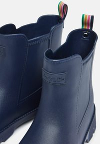 Mornaričke gumene čizme do gležnja s glatkom završnom obradom, s uzdignutim logotipom, valovitim otvaranjem i višebojnom trakastom petljom.