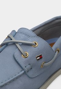 Φως μπλε καστόρινο παπούτσι τύπου boat με γκρι κορδόνια, χρυσά οπτικά και μικρό έμβλημα σημαίας κόκκινου, λευκού και μπλε στο πλάι.