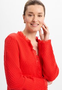 Roter strukturierter Cardigan mit fransigen Kanten, V-Ausschnitt und langen Ärmeln. Der Strickstoff hat ein dezentes offenes Muster.