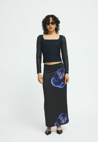 Schwarz-blumiger Maxirock mit großem blauen Blumenmuster, kombiniert mit einem taillierten, dunklen Spitzenoberteil mit langen Ärmeln und quadratischem Ausschnitt.