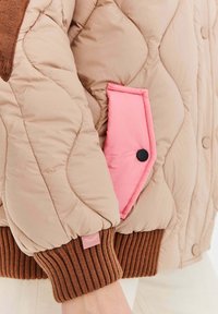 Přebíraná béžová bunda s růžovou kapsou na tlakové knoflíky a hnědými žebrovanými manžetami. Měkká textura s ostrým, polstrovaným designem.