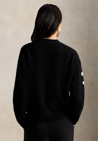 Mujer con cabello negro liso, vestida con un suéter negro con puntos blancos en la manga, de pie frente a un fondo beige neutro.