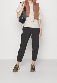 Person som bär en brun vadderad väst, beige kortärmad skjorta, svarta cargo-byxor, gröna sneakers och håller en svart axelväska.