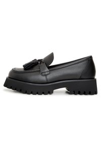 Mocassino in pelle nera con dettaglio in frange, caratterizzato da una suola spessa e scanalata e un design slanciato. Texture liscia con cuciture dettagliate.