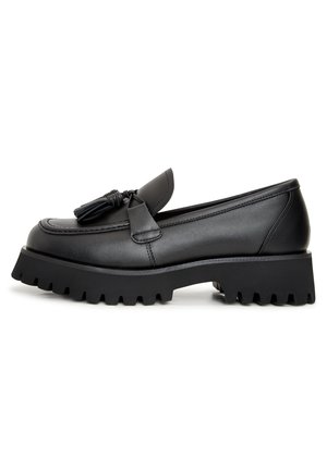Mocassin en cuir noir avec un accent à franges, présentant une semelle épaisse et nervurée ainsi qu'un design épuré. Texture lisse avec des coutures détaillées.