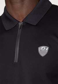 Juodas polo marškinėlis su užtrauktuku ant apykaklės, turintis logotipo aplikaciją su dryžuotu raštu. Medžiaga atrodo lygus ir pralaidus orui.