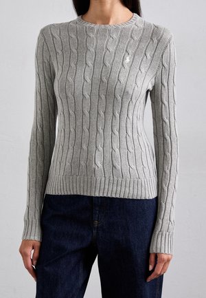 Femme portant un pull cintré gris clair en maille torsadée à manches longues et un jean bleu foncé, debout devant un fond clair uni.