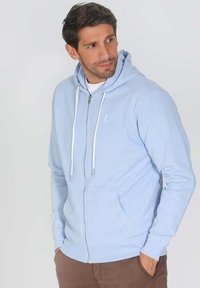 Haut de sweatshirt zippé bleu clair en tissu doux, avec une poche avant, des cordons et des manches côtelées. Petit logo sur la poitrine.