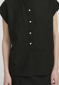 Camicia nera senza maniche con cinque bottoni bianchi sulla parte anteriore, indossata da una persona con le braccia visibili ai lati.