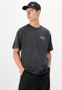 Young man wearing a black Nike cap, faded black Nike SB t-shirt, and black pants, looking to the side with hands in pockets.