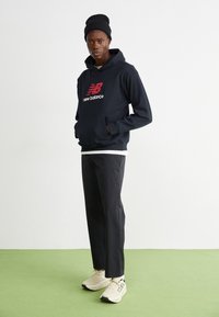 Schwarzer Hoodie mit rotem Logo, schwarze Hose und beiger Sneaker. Das Outfit zeichnet sich durch eine entspannte Passform und minimalistische Designelemente aus.