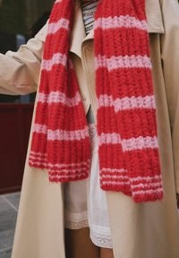 Person med beige frakke og stribet strikscarf i pink og rød over en mønstret sweater og hvid nederdel.