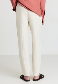 Persona che indossa pantaloni dritti color crema, una camicia larga a maniche lunghe color corallo e sandali neri intrecciati, in piedi su un pavimento chiaro.