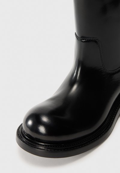 Svart läderankleboot med en glansig yta, rund tå, kontrasterande sömmar och en tjock gummisula. Sidoseamdetalj synlig.