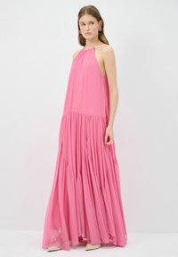 Vestido maxi rosa fluido con escote halter, textura plisada y diseño en capas. La modelo está de pie con tacones claros y pendientes.