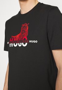 T-shirt en coton noir avec un motif de tigre rouge et un texte en gras "HUGO" en blanc et rouge, doté d'un col rond et de manches courtes.
