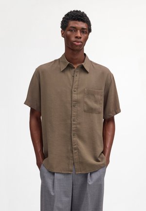 Junger Mann mit kurzen lockigen Haaren trägt ein braunes kurzärmeliges Hemd mit Brusttasche und graue Plissee-Hose, Hände in den Taschen.