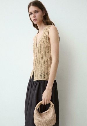 Giovane donna con top beige senza maniche in maglia e gonna nera che tiene una borsa beige intrecciata su uno sfondo bianco uniforme.